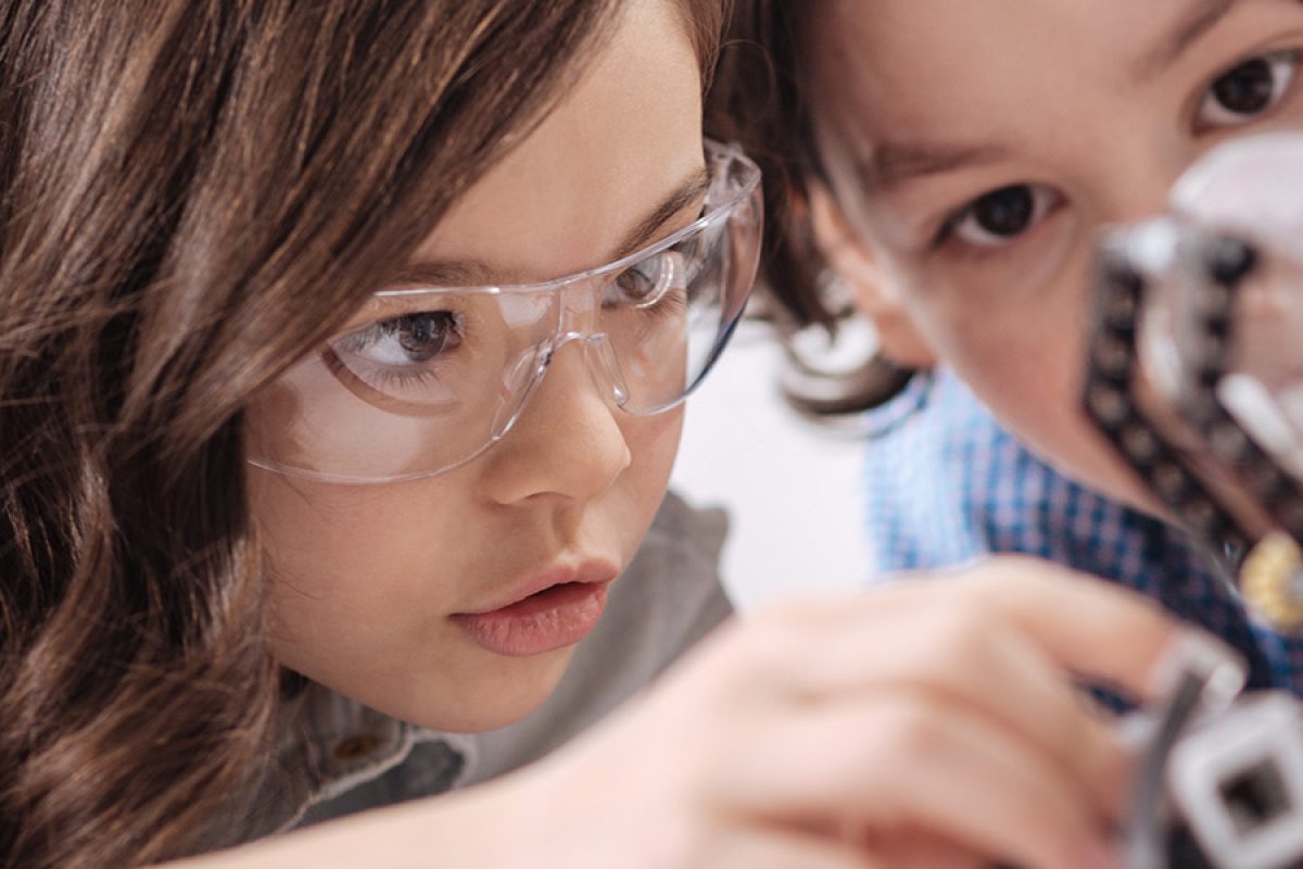 Zwei Kinder bauen konzentriert an einem Roboter. Eines trägt eine Schutzbrille.