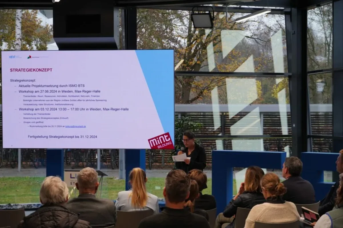 Eine Frau präsentiert vor Publikum ein Strategiekonzept auf großer Leinwand in heller Veranstaltungshalle.