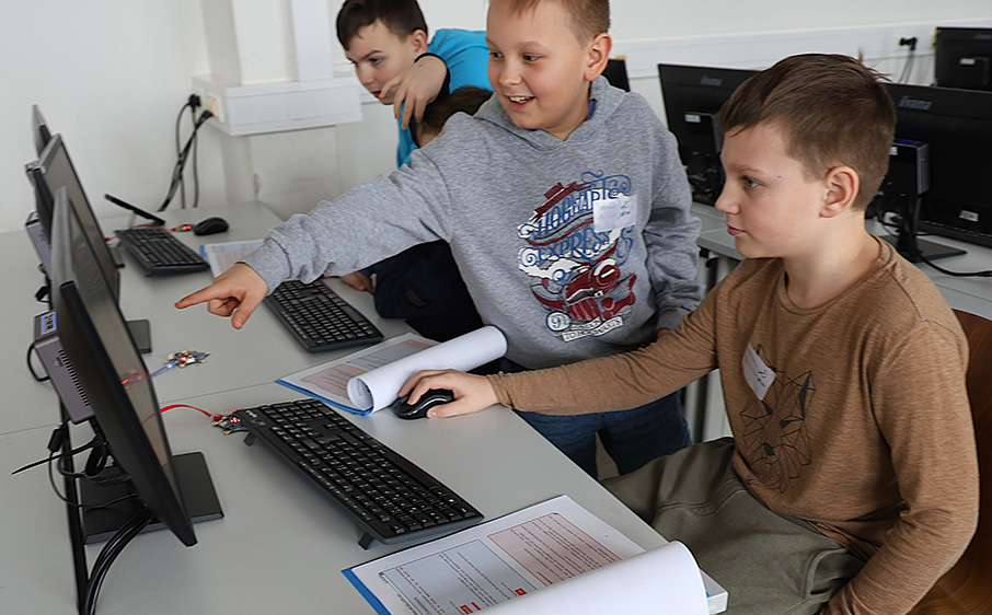 Drei Jungen arbeiten gemeinsam an einem Computer und zeigen lachend auf den Bildschirm.