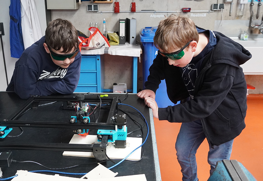 Zwei Jungen beobachten mit Schutzbrillen eine Lasergravur-Maschine bei der Arbeit.