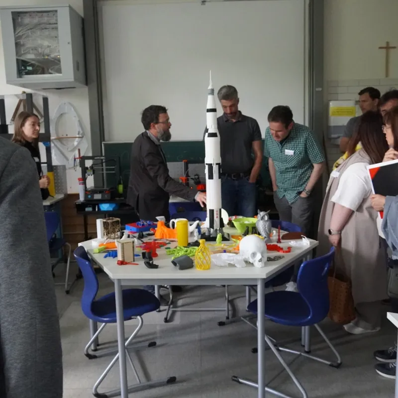 Erwachsene betrachten 3D-gedruckte Objekte, darunter eine große Rakete, in einem Technikraum.