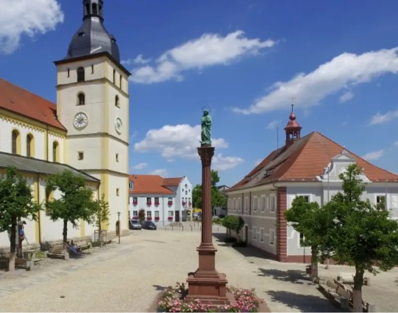 Der Stadtplatz Tirschenreuths zeigt Kirche, Marienstatue, barocke Bauten und Blumen bei Sonnenschein.